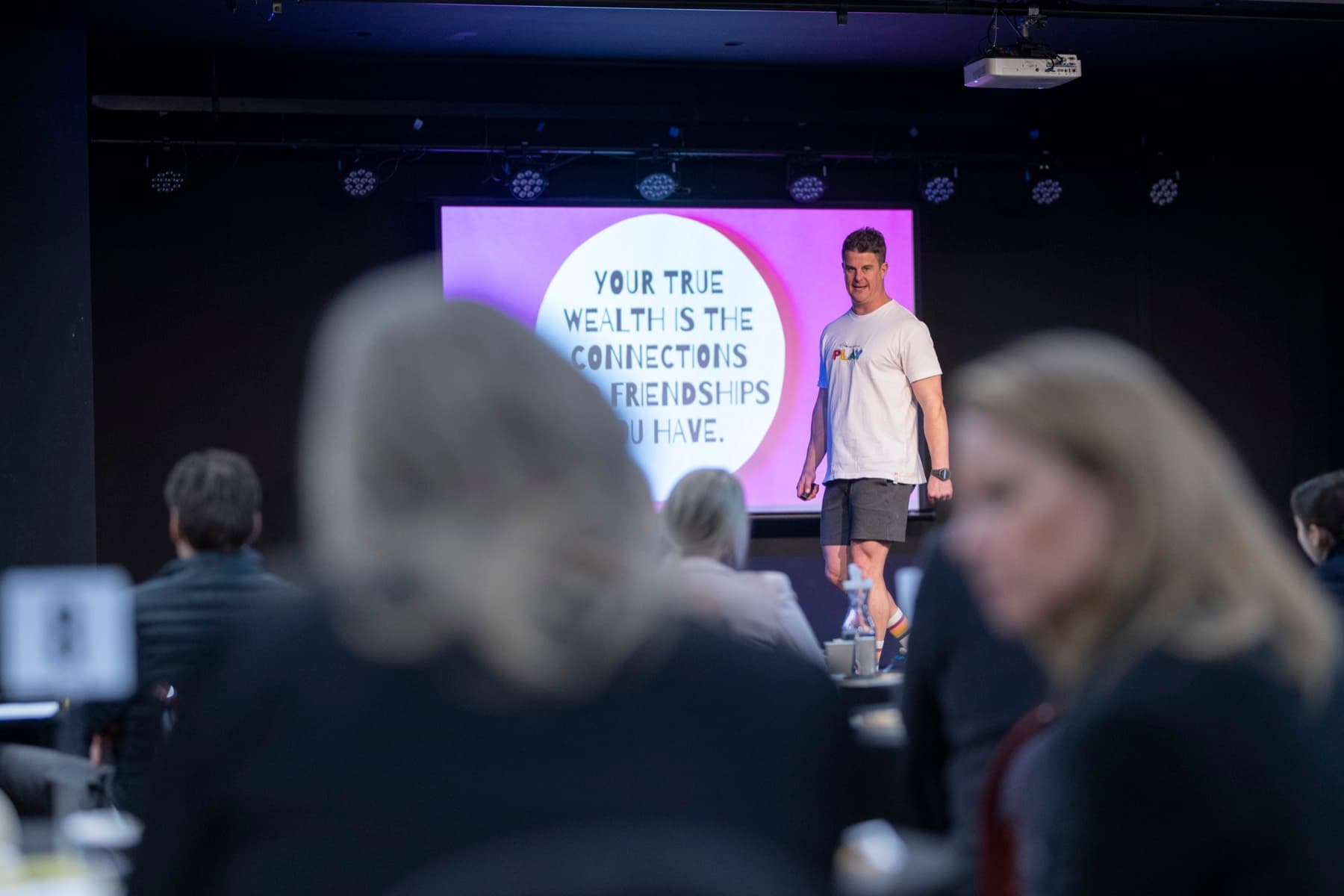 Dale on stage in front of a slide reading “your true wealth is the connections and friendships you have”