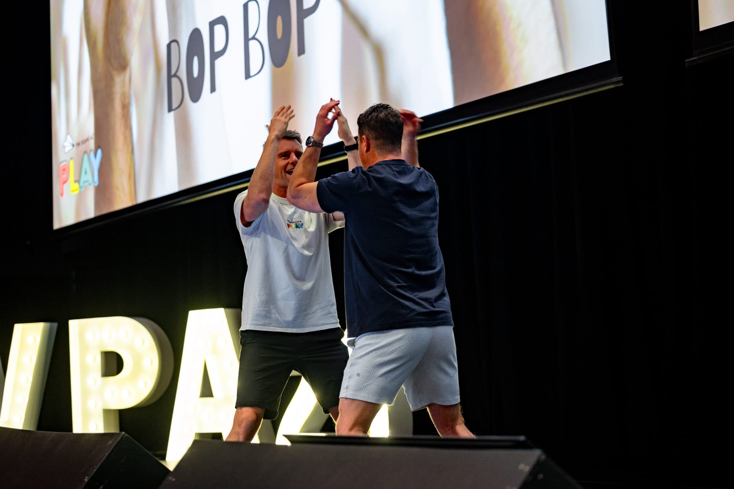 Two presenters high-fiving on stage under a big screen and lit-up marquee letters